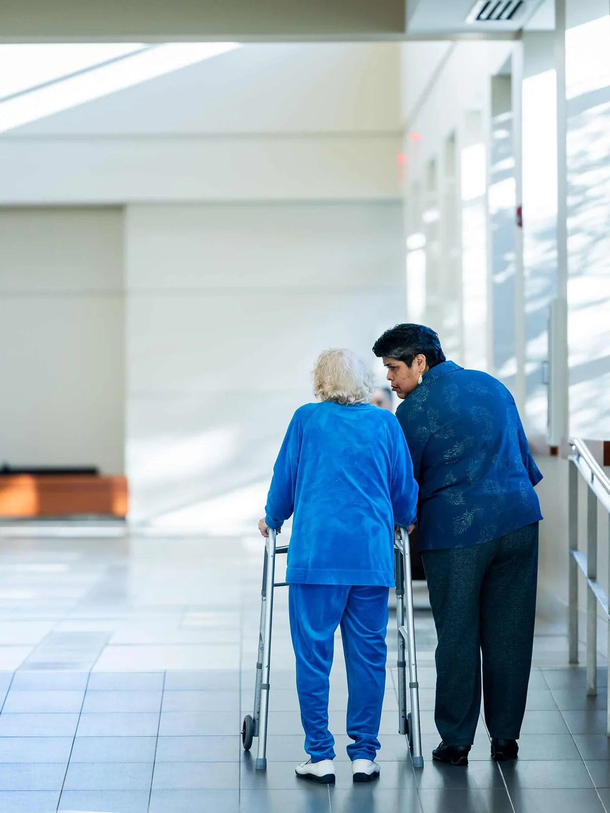 Senior living resident with a walker and a caregiver in a sunlit hallway